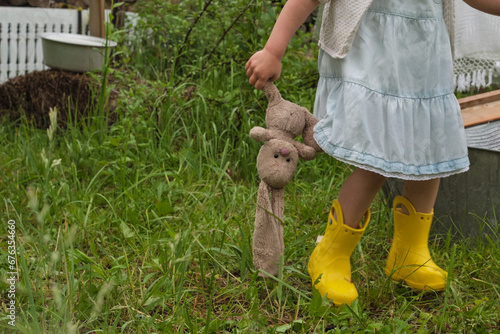 Child in countryside setting holding a cherished stuffed toy, evoking themes of innocence, sustainable living, and reconnecting with nature. Yellow boots highlight a playful contrast. photo