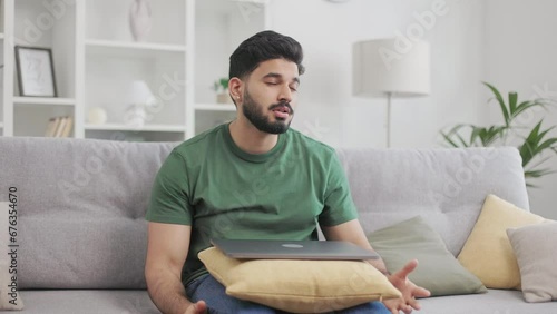 Upset indian guy spreading hands in disappointment and sigh heavily because of getting bad news. Handsome male freelancer sitting on comfy couch and closing wireless laptop on knees.