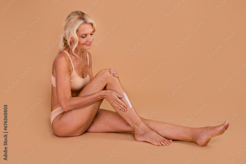 Full length profile photo of lovely skinny aged lady sit floor apply cream silky legs isolated on beige color background Full length profile photo of lovely skinny aged lady sit floor apply cream silky legs isolated on beige color background