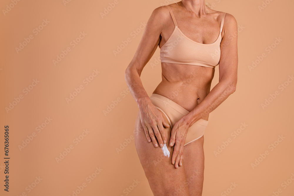 Photo of sexy slender senior lady dressed lingerie applying legs anti cellulite cream empty space isolated beige color background