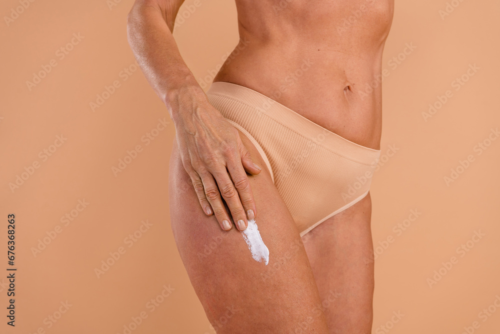Photo of slender tanned senior lady dressed lingerie applying legs anti cellulite cream empty space isolated beige color background Photo of slender tanned senior lady dressed lingerie applying legs anti cellulite cream empty space isolated beige color background