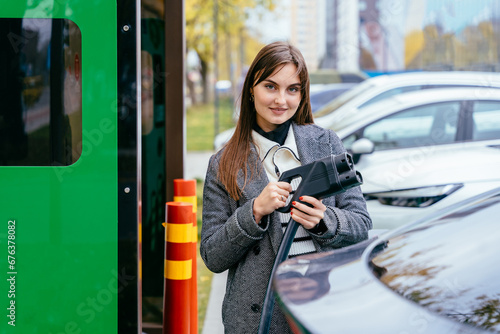 Portrait of young woman holding electric plug while waiting to charge an electric car at a city public charger station. Electric car concept. Green travelling.