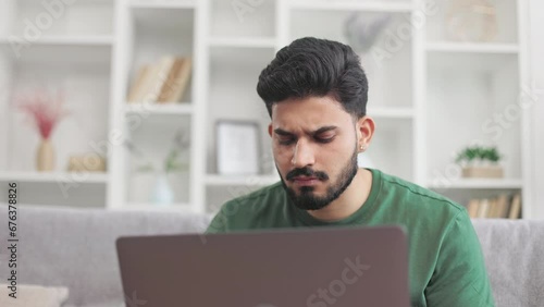 Casually dressed young guy scratching bearded chin while looking on laptop screen with pensive facial expression. Indian man sitting on couch in doubt during online work at home. photo
