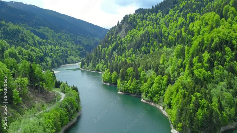 4k aerial flight Petrimanu Lake and a gravel road winding along its shores. Spring time, the trees are blooming. The clouds are reflecting into the lake's waters. Carpathia, Romania. 4k aerial flight Petrimanu Lake and a gravel road winding along its shores. Spring time, the trees are blooming. The clouds are reflecting into the lake's waters. Carpathia, Romania.