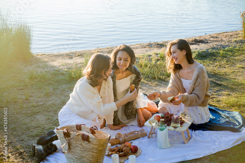 Young beautiful women of 25 years old on an autumn picnic near the lake. Glass of white wine, pastries. Happy models chatting merrily. Sunset. © Kristina89