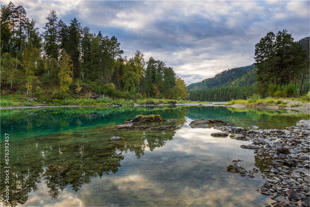 Obraz premium Idyllic view of calm lake and rocky shore with autumn trees in background against cloudy sky at Altai, Russia 