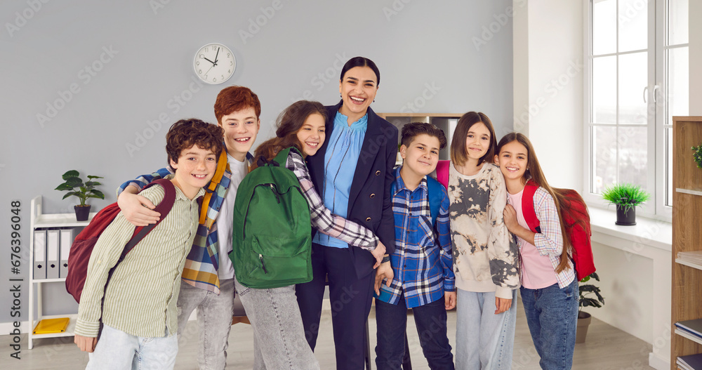 Portrait of a happy schoolchildren group with backpacks on their shoulders standing in a row with their teacher meeting after holidays in classroom at school and hugging. Back to school concept. Portrait of a happy schoolchildren group with backpacks on their shoulders standing in a row with their teacher meeting after holidays in classroom at school and hugging. Back to school concept.