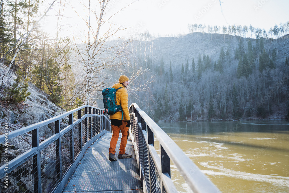 Trekking in nature in a mountainous area with a backpack on your back, late autumn, the first frosts, a man walking on an iron bridge along a pond.