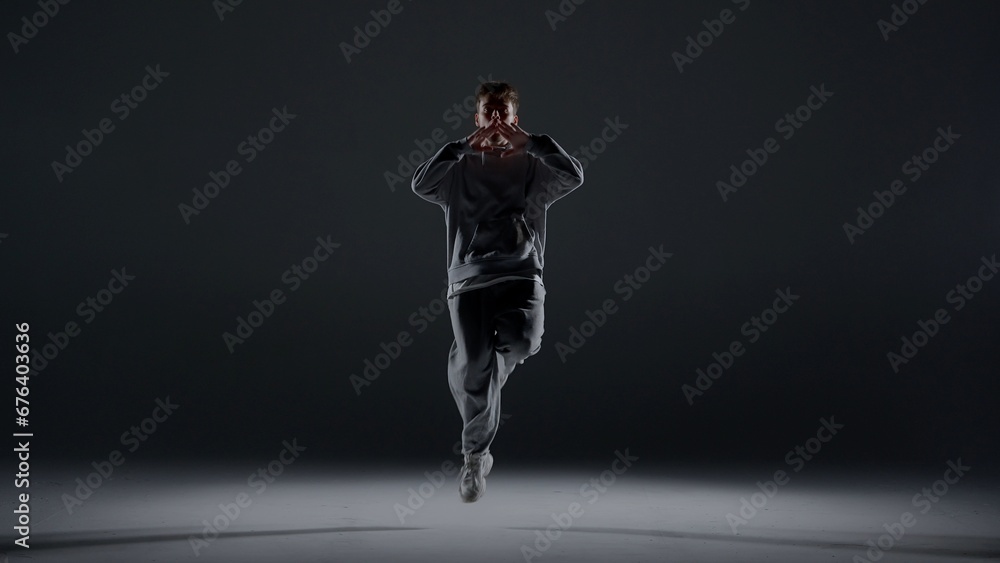 A young man stands against a dark background. He is highlighted by a white light from below. Creates a light haze. Demonstrates a dance movement by bouncing on one foot. He is plastic, rhythmic