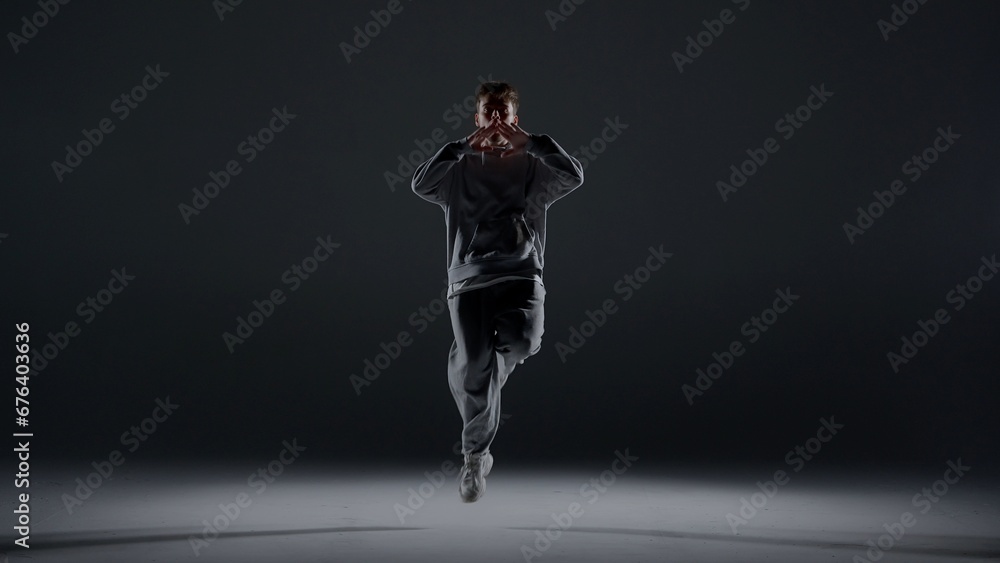A young man stands against a dark background. He is highlighted by a white light from below. Creates a light haze. Demonstrates a dance movement by bouncing on one foot. He is plastic, rhythmic A young man stands against a dark background. He is highlighted by a white light from below. Creates a light haze. Demonstrates a dance movement by bouncing on one foot. He is plastic, rhythmic