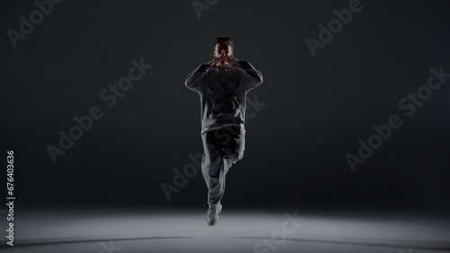 A young man stands against a dark background. He is highlighted by a white light from below. Creates a light haze. Demonstrates a dance movement by bouncing on one foot. He is plastic, rhythmic