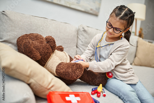 happy kid in casual wear and eyeglasses playing doctor with teddy bear on sofa in living room photo