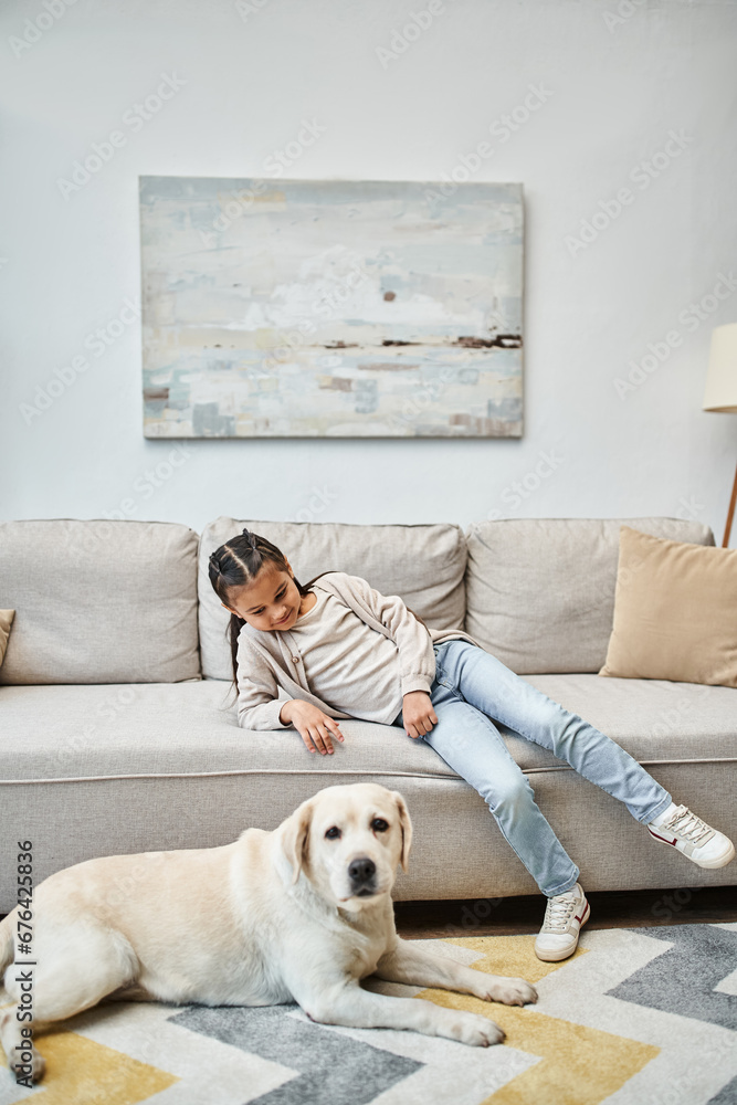 cute girl smiling and sitting on sofa while looking at labrador in modern living room, kid and dog cute girl smiling and sitting on sofa while looking at labrador in modern living room, kid and dog