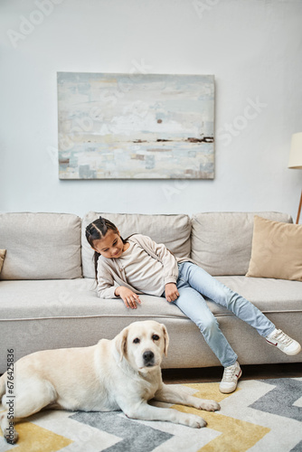 cute girl smiling and sitting on sofa while looking at labrador in modern living room, kid and dog