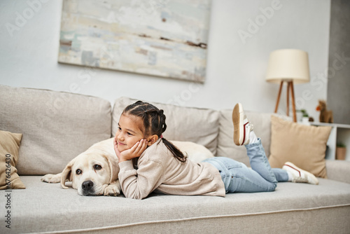 happy girl smiling and lying on sofa with cute labrador in modern living room, pet and child