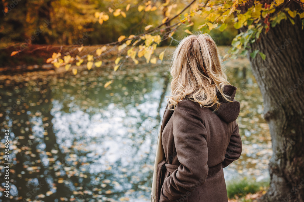 Pensive woman standing at lake in autumn park. Lonely sad female person looking at reflection on water surface