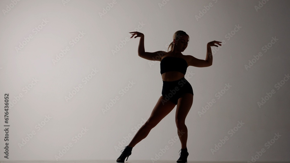 The frame is on a gray background. The woman is slightly darkened. She is dressed in open clothes and high heels. Demonstrates dance movement, posture, legs and arms apart. She is sexy, plastic