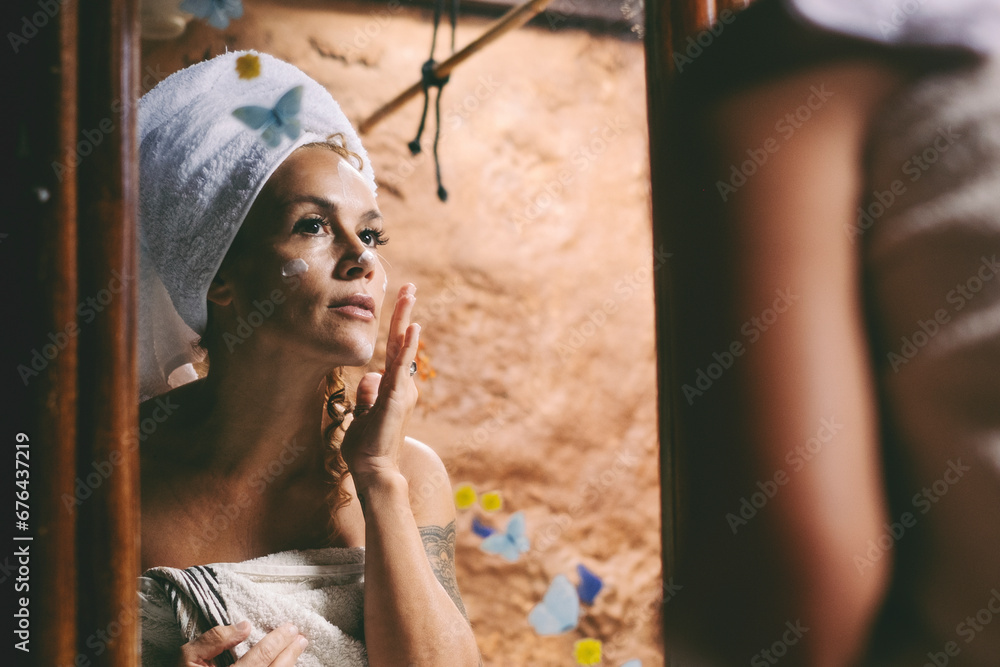 Adult attractive woman use beauty cream on her face looking in the mirror at home. Skin care anti aging treatment in the bathroom. Female people with white towel on head after wash Adult attractive woman use beauty cream on her face looking in the mirror at home. Skin care anti aging treatment in the bathroom. Female people with white towel on head after wash
