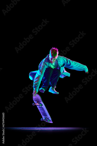 Dynamic image of young man in casual clothes in motion, training, skateboarding against black studio background in neon light. Concept of professional sport, competition, training, action.