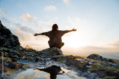 Girl hugging the rays of the morning sun in the mountains sitting on a stone, the sunrise in the mountains, the view from the back of a man on the top of the mountain, the morning light. © Aleksey