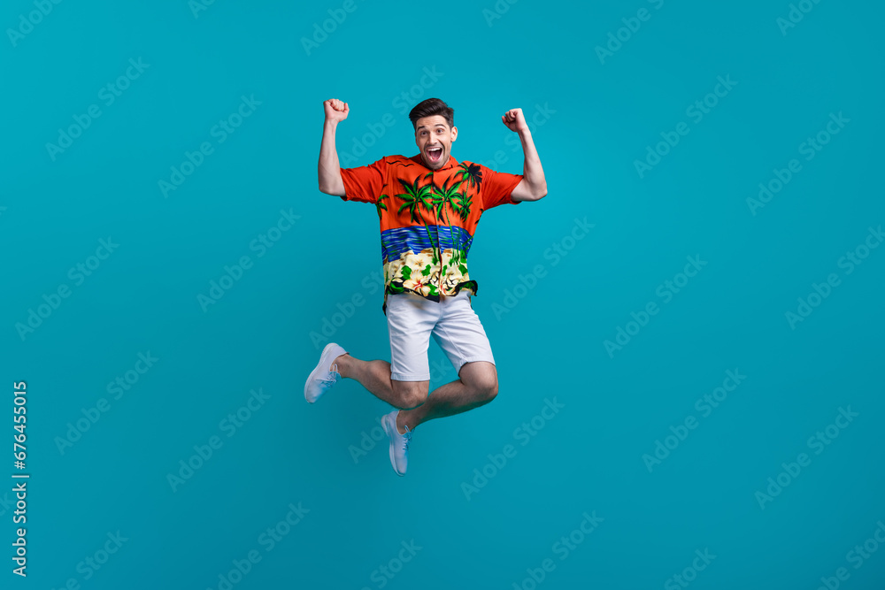 Full body photo of funky jumping crazy guy fists wear tropics shirt celebrate summertime season hooray isolated on blue color background Full body photo of funky jumping crazy guy fists wear tropics shirt celebrate summertime season hooray isolated on blue color background