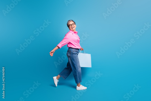 Full size photo of carefree positive person with grey hair go shopping in empty space hold new clothes isolated on blue color background