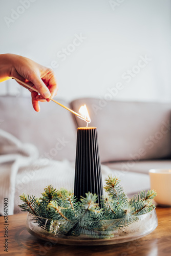 Female hand lighting a candle with a match in winter cozy composition for hygge home mood. Black candle and spruce branches on the coffee table in living room. Natural Christmas decorations for home. photo