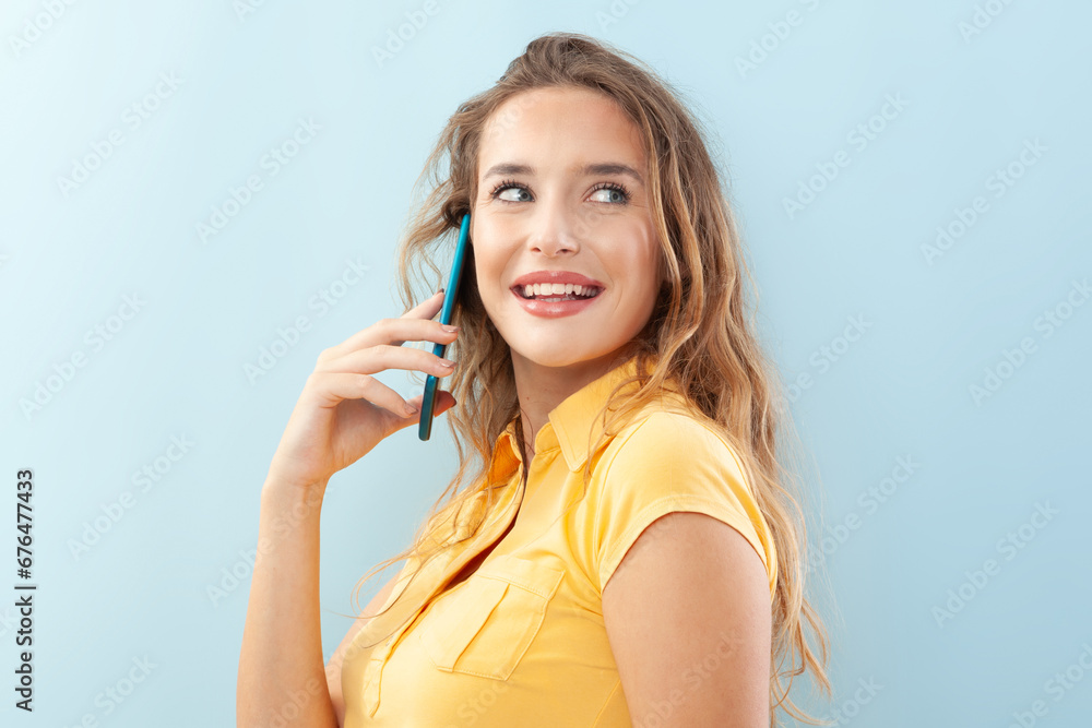 Cheerful and beautiful young woman talking on cell phone, with long hair wearing a yellow blouse. Isolated on light blue background, portrait of laughing girl with big blue eyes she looks to the side. Cheerful and beautiful young woman talking on cell phone, with long hair wearing a yellow blouse. Isolated on light blue background, portrait of laughing girl with big blue eyes she looks to the side.