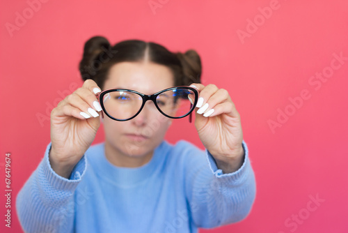 a young pretty woman in a blue sweater with poor eyesight holds glasses in her hands, squints and looks through them into the camera. Vision problems. Ophthalmology photo