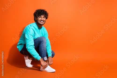 Full length photo of cheerful indian guy advertising new sneakers adidas brand lacing up sitting floor isolated on orange color background photo