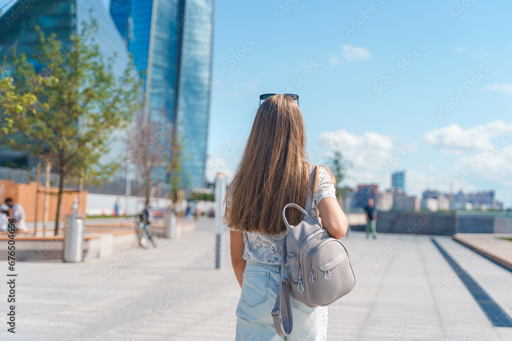 A beautiful young girl in jeans with long hair with a backpack walks down the street with a view of a high-rise building in St. Petersburg
