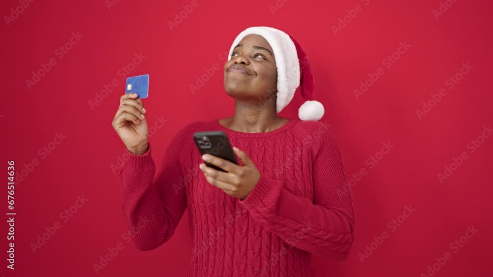 African american woman using smartphone and credit card wearing christmas hat over isolated red background African american woman using smartphone and credit card wearing christmas hat over isolated red background