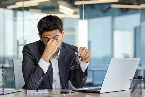 Overtired and overwhelmed businessman at workplace inside office, man took off glasses rubbing eyes, dizziness migraine and headache, man in business suit working late with laptop at workplace.