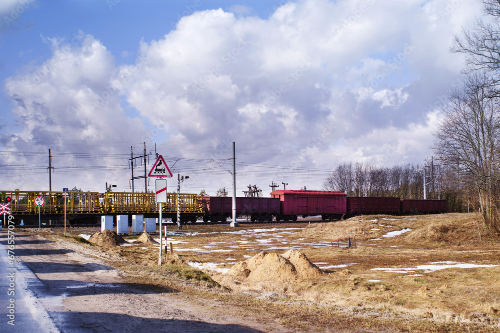 A container freight train passes by a railway crossing against a background of a cloudy sky on a sunny spring day. Freight transportation.