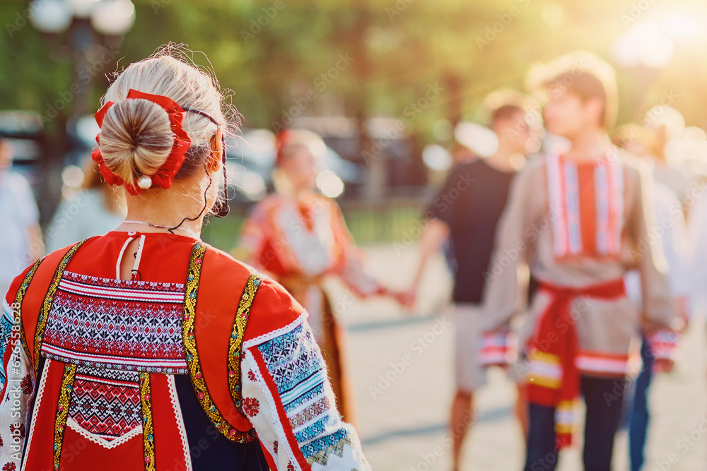 A girl in a Russian national costume from the back. A folk ensemble dances on the embankment. Bright sunlight. World Folklore Day concept.