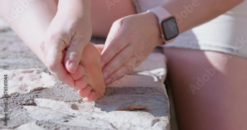 Woman in denim shorts sits on concrete bench, clears dirt from her bare feet, carefully shakes off her legs, checks her fingers for splinters Leg discomfort, calluses, chafing Examination for injurie photo