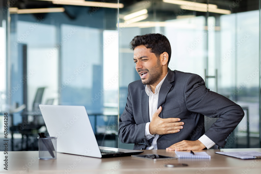 Chest pain, heart attack in businessman at workplace, man holding hands on chest, breathing hard, boss in business suit working inside office with laptop. Chest pain, heart attack in businessman at workplace, man holding hands on chest, breathing hard, boss in business suit working inside office with laptop.