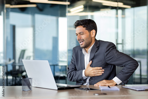 Chest pain, heart attack in businessman at workplace, man holding hands on chest, breathing hard, boss in business suit working inside office with laptop. photo