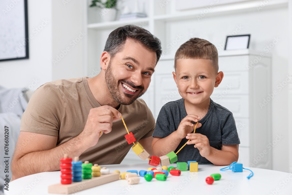 Motor skills development. Father and his son playing with wooden pieces and string for threading activity at table indoors Motor skills development. Father and his son playing with wooden pieces and string for threading activity at table indoors