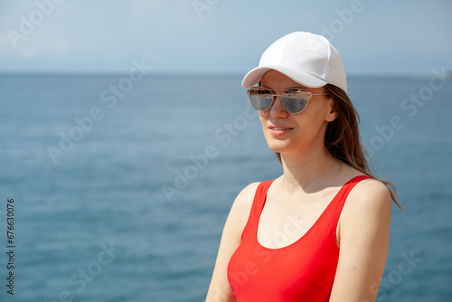 A beautiful girl in a red swimsuit with long hair  glasses and a white cap looks at the blue sea.