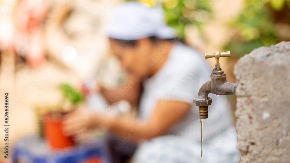 Close-up of a water tap with a smiling woman enjoying the plants in the vegetable garden in the background. Sustainable urban agriculture concept. Selective focus.