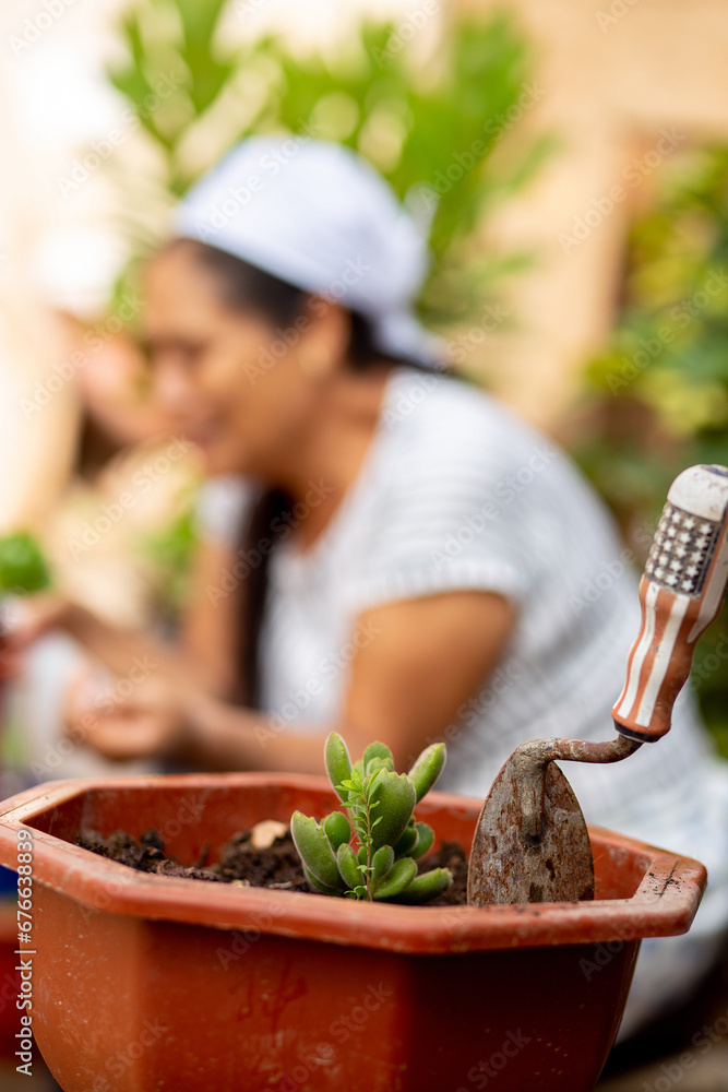 Close-up of a cultivation tool in the foreground, with a blurred background of a smiling woman enjoying the plants in the vegetable garden. Sustainable urban agriculture concept. Selective focus.