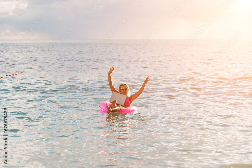 Woman works on laptop in sea. Freelancer, blond woman in sunglases floating on an inflatable big pink donut with a laptop in the sea at sunset. People summer vacation rest lifestyle concept.