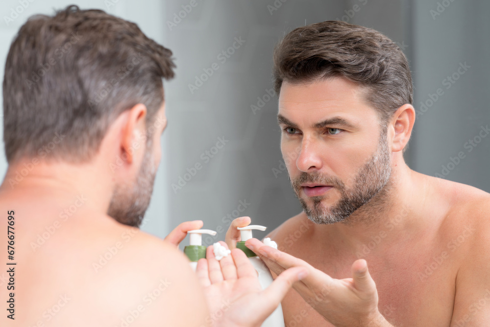 Fototapeta premium Portrait of millennial man applying cream on face skin. Handsome serious man enjoy fresh perfect skin, applying cream. Portrait of beauty man with cosmetic cream on skin. Facial treatment. Male skin