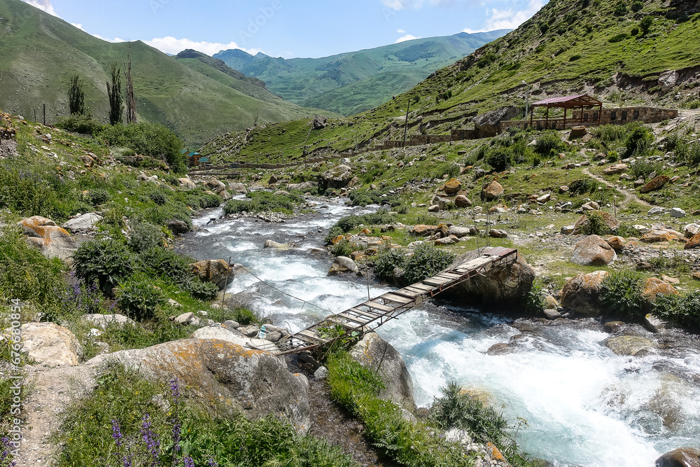 Suspension bridge over the mountain river. The picturesque gorge of the Jylgy-su river. The Caucasus Mountains near the ancient city of Eltuby. Next to the grotto with an ancient human parking lot. Suspension bridge over the mountain river. The picturesque gorge of the Jylgy-su river. The Caucasus Mountains near the ancient city of Eltuby. Next to the grotto with an ancient human parking lot.