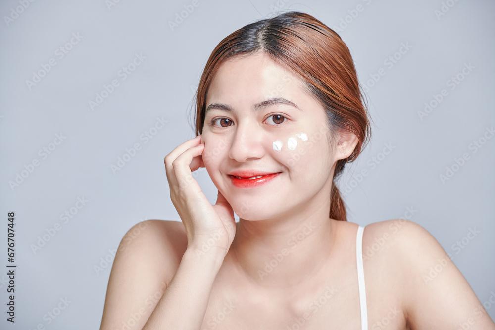 Positive young female in white top applying moisturizing cream on face. Close up beauty portrait of laughing beautiful half naked woman applying face cream and looking away isolated grey background. Positive young female in white top applying moisturizing cream on face. Close up beauty portrait of laughing beautiful half naked woman applying face cream and looking away isolated grey background.
