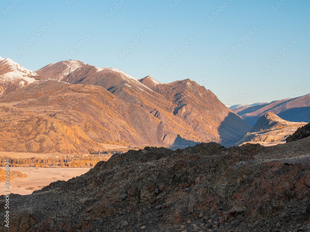 Autumn snow mountains at noon. Bright mountain landscape with a snowy rock in golden sunlight. Natural background of a walk through the rocky mountains with sharp rocks and blue sky.