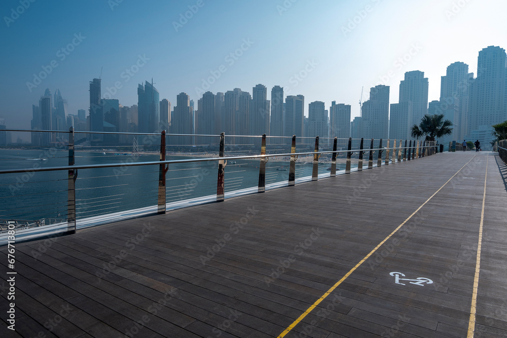 Dubai city silhouette and sea behind the timbered walking terrace bicycle lanes in Dubai blue waters island daylight misty hazy skies. Dubai city silhouette and sea behind the timbered walking terrace bicycle lanes in Dubai blue waters island daylight misty hazy skies.