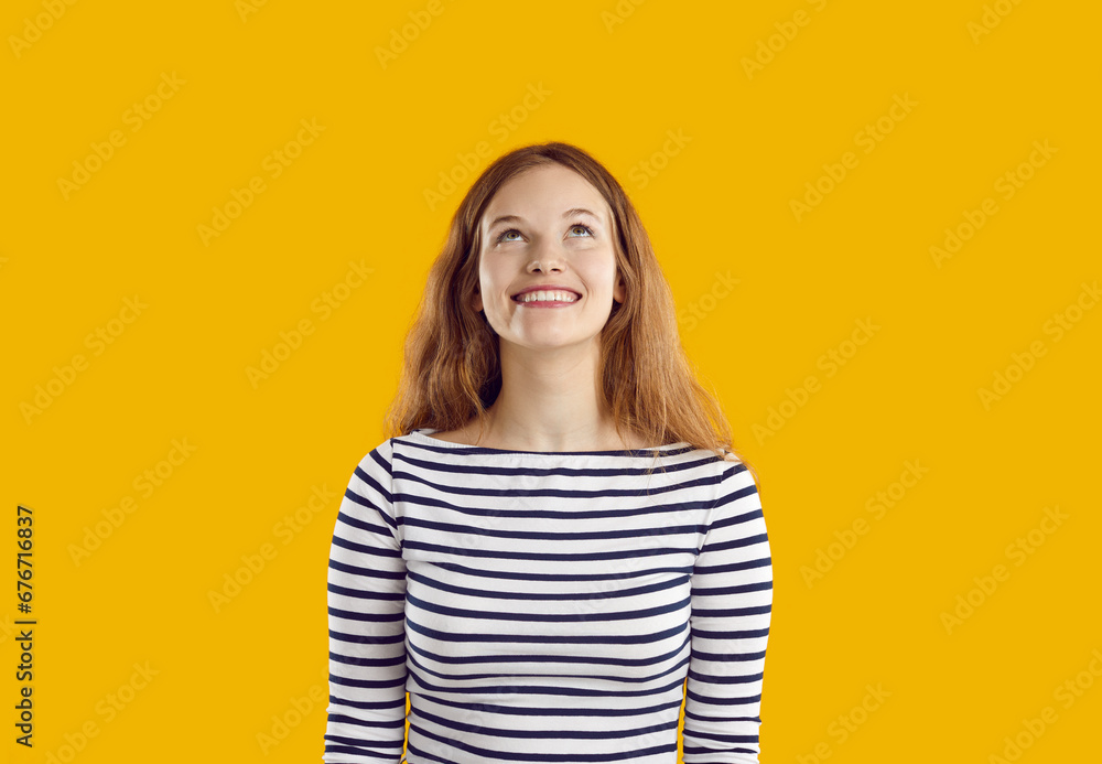 Young cheerful long haired Caucasian woman millennial in striped clothes smiling broadly looks up and rejoices at seen advertising offer stands on orange studio background. Copy space Young cheerful long haired Caucasian woman millennial in striped clothes smiling broadly looks up and rejoices at seen advertising offer stands on orange studio background. Copy space