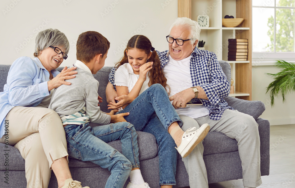 Happy grandparents and children having fun on the weekend. Funny, cheerful grandmother and grandfather tickling happy little kids grandchildren while sitting on the sofa at home all together Happy grandparents and children having fun on the weekend. Funny, cheerful grandmother and grandfather tickling happy little kids grandchildren while sitting on the sofa at home all together